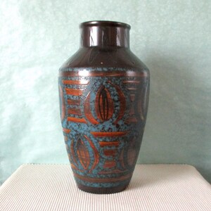 Puede incluir: Un jarrón de cerámica marrón alto con una superficie texturizada y un cuello estrecho. El jarrón presenta un patrón de formas geométricas naranjas y azules. El jarrón mide aproximadamente 30 cm de alto.