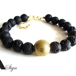 Könnte beinhalten: Ein Armband aus schwarzen Lavasteinen mit einem goldenen Perlen in der Mitte. Das Armband hat einen goldenen Verschluss.