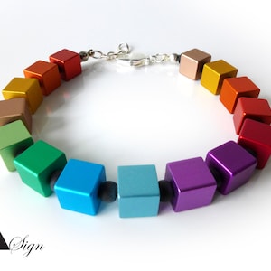 Op de afbeelding: Een kleurrijke armband met vierkante kralen in regenboogkleuren, waaronder rood, oranje, geel, groen, blauw en paars. De kralen zijn verbonden door een zilveren ketting en zwarte afstandhouders. Een levendig accessoire.