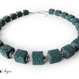 Op de afbeelding: Een ketting met een reeks kubusvormige, poreuze lavasteenkralen in een blauwgroene kleur, afgewisseld met kleine, gladde, metallic zilverkleurige tussenkralen. De ketting is voorzien van een zilverkleurige metalen sluiting en de tekst A Sign.