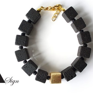 Peut inclure: Un bracelet en pierre de lave noire avec des accents dorés. Le bracelet est composé de perles carrées et possède un fermoir doré.