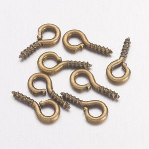 Può includere: Un gruppo di dieci ganci ad occhiello a vite in oro antico. I ganci sono in metallo e hanno un piccolo anello nella parte superiore. Vengono utilizzati per appendere oggetti.