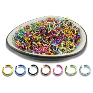 Könnte beinhalten: Eine Keramikschale gefüllt mit einer Sammlung farbenfroher Sprungringe in verschiedenen Metallic-Ausführungen. Darunter eine Reihe einzelner Sprungringe in Gold, Blau, Schwarz, Kupfer, Pink, Grün und Hellblau, die die Farboptionen zeigen.