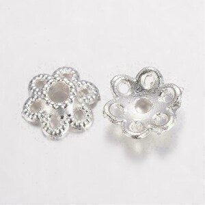 Puede incluir: Dos componentes de joyería plateados con forma de flor. Cada componente tiene un agujero central rodeado de seis pétalos, cada uno con un agujero más pequeño. Los componentes son de metal brillante.