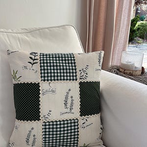Può includere: Cuscino decorativo quadrato con un design patchwork. Il cuscino presenta quadrati di tessuto bianco con illustrazioni botaniche e testo, motivo a quadri verde e pois verdi. Il cuscino è su un divano bianco.