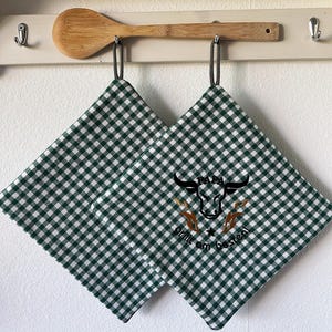 Può includere: Due presine a quadretti verdi e bianchi con una grafica di toro e testo, appese a un ripiano bianco con un cucchiaio di legno. Le presine hanno un anello per appenderle.