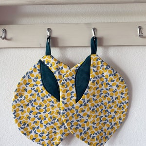 Könnte beinhalten: Zwei Topflappen mit Zitronenmuster und grünen Stoffakzenten, die an Haken hängen. Die Topflappen sind weiß mit einem Muster aus gelben Zitronen und blauen Blättern.