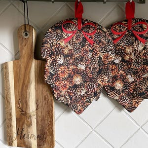 Könnte beinhalten: Ein Holzschneidebrett mit einem Hirschkopf-Design und dem Wort "Heimat" wird neben zwei blattförmigen Topflappen angezeigt. Die Topflappen haben ein Tannenzapfenmuster und rote Schleifenakzente.