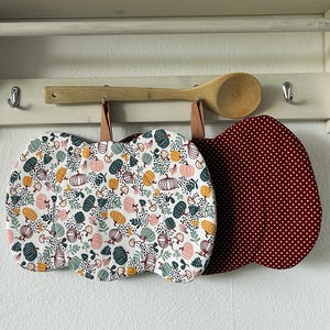 Könnte beinhalten: Zwei kürbisförmige Topflappen aus Stoff, die an einem Holzregal hängen. Einer hat einen weißen Hintergrund mit einem Muster aus kleinen Kürbissen, Pilzen und Blättern in Orange, Rosa und Dunkelgrün. Der andere ist dunkelrot mit weißen Punkten.