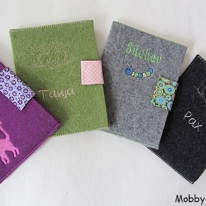 Könnte beinhalten: Vier Filz-Buchhüllen in Lila, Grün, Grau und Schwarz. Jede Hülle hat ein einzigartiges Design, einschließlich gesticktem Text und aufgenähten Patches. Die lila Hülle zeigt eine Katzen-Silhouette, während die grüne Hülle den Namen "Tanja" trägt. Die graue Hülle sagt "Bücher".