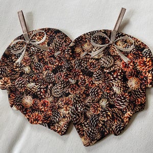 Può includere: Due presine in tessuto a forma di foglia con un motivo ripetuto di pigne nei toni del marrone, beige e nero. Ogni presina ha un piccolo fiocco di nastro e un anello per appenderla. Le presine sono larghe circa 20 cm.