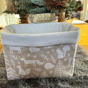 Könnte beinhalten: Ein rechteckiger Aufbewahrungskorb mit weißem Innenraum und beigefarbenem Außenbereich. Das Äußere zeigt ein sich wiederholendes Muster aus weißen Waldtieren, Bäumen und Sternen. Der Korb steht auf einer strukturierten, grauen Oberfläche.