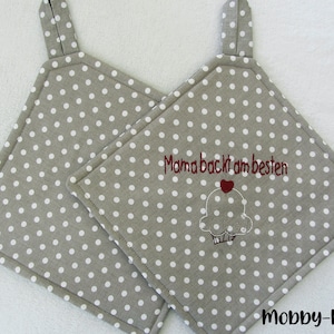 Può includere: Due presine grigie con pois bianchi. Una presina ha il testo "Mama backt am besten" ricamato con un cuore rosso e un'immagine di cupcake.