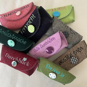 Peut inclure: Plusieurs étuis à lunettes en feutre de différentes couleurs, dont rose, vert et violet. Chaque étui est orné de texte brodé et d'un bouton décoratif. Conçus pour protéger les lunettes.