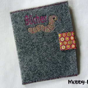 Könnte beinhalten: Grauer Filzbuchumschlag mit rosa Nähten und dem in rosa gestickten Wort "Bücher". Ein rosa-gelber Stoff-Patch mit geometrischem Muster ist am Umschlag befestigt.