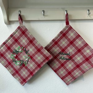 Könnte beinhalten: Zwei quadratische Topflappen aus rot-weiß-grün kariertem Stoff. Einer ist mit einem gestickten grünen Kranz mit weißen Beeren verziert, während der andere den gestickten grünen Schriftzug "Advent" mit drei Punkten zeigt.