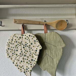 Könnte beinhalten: Zwei blattförmige, gesteppte Topflappen, die an einem Holzlöffel hängen. Ein Topflappen hat einen beigen Hintergrund mit einem kleinen grün-weißen Blumenmuster, der andere ist olivgrün mit dünnen, vertikalen weißen Streifen.