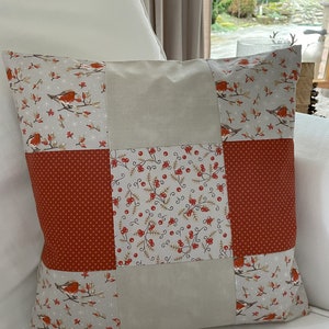 Peut inclure: Un coussin patchwork avec un fond beige et des accents rouges. Le coussin est composé de quatre tissus différents, chacun avec un motif différent. Les motifs comprennent un petit pois rouge, un motif floral avec des baies rouges et un motif avec de petits oiseaux sur des branches. Le coussin est posé sur un canapé blanc.