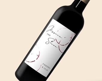 May include: A white wine bottle label with a handwritten inscription that reads "Jessica and Steve" and "Cheers! 8.14.22". The label has a burgundy wine stain design.