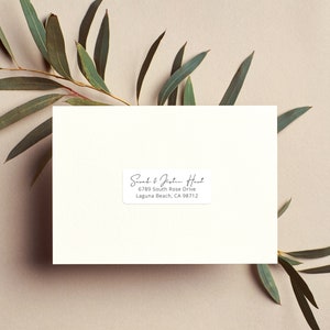 Puede incluir: Una etiqueta rectangular blanquecina con texto negro, con los nombres "Sarah & Joshua Hart" y una dirección. La etiqueta está sobre un fondo neutro con hojas verdes de eucalipto.