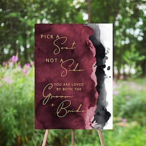 May include: A burgundy and black watercolor wedding sign with gold lettering that reads "Pick a Seat Not a Side. You are loved by both the Groom + Bride."