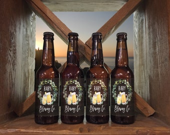 May include: Four brown glass beer bottles with labels that say "Baby Brewing Co." and feature a watercolor illustration of a beer mug and greenery. The bottles are arranged in a row on a wooden shelf with a sunset view in the background.