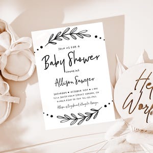 May include: A white baby shower invitation with black calligraphy text. The invitation reads "Join us for a Baby Shower honoring Allison Sawyer" and includes the date, time, and location of the event. The invitation is decorated with simple black floral illustrations.