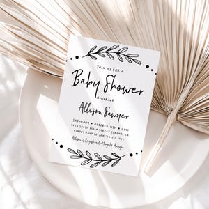 May include: A white baby shower invitation with black calligraphy text. The text reads "Join us for a Baby Shower honoring Allison Sawyer" and includes the date, time, and location of the event. The invitation is decorated with simple black floral illustrations.