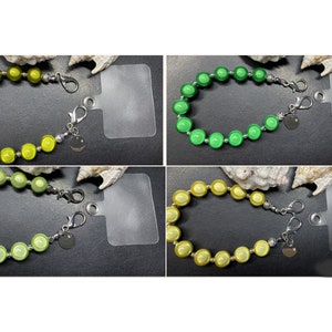 Könnte beinhalten: Vier Armbänder mit Perlen in Grün- und Gelbtönen. Jedes Armband hat runde Perlen, silberne Akzente und einen Karabinerverschluss. An jedem Verschluss hängt ein kleiner, runder Anhänger. Die Armbänder werden auf einer schwarzen Oberfläche präsentiert.