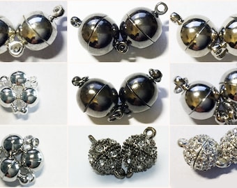 May include: A collection of nine magnetic jewelry clasps in various finishes including polished silver, gunmetal, and crystal-encrusted spheres. Each clasp features two spherical components with small loops for attachment to necklaces or bracelets.