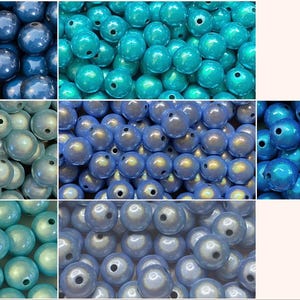Könnte beinhalten: Eine Sammlung runder, kugelförmiger Perlen in verschiedenen Blau- und Türkistönen mit irisierenden Perlmutt-Finishes. Jede Perle hat ein mittiges Loch zum Auffädeln. Das Sortiment umfasst dunkelblaue, leuchtend türkise, hellblaue und perlmuttfarbene Töne.