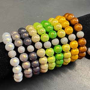 Può includere: Una collezione di braccialetti di perline in una sfumatura di colori, dal grigio chiaro al marrone. Ogni braccialetto presenta grandi perline rotonde ed è intervallato da perline di accento argentate con strass. I braccialetti sono esposti su una superficie nera.