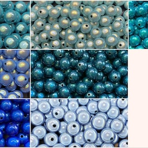 Könnte beinhalten: Eine Sammlung schillernder Perlen in verschiedenen Blau- und Türkistönen. Die Perlen sind rund mit glänzender Oberfläche und einem kleinen Loch zum Auffädeln. Das Bild zeigt eine Vielzahl von Farben, darunter Hellblau, Dunkelblau und Türkis.