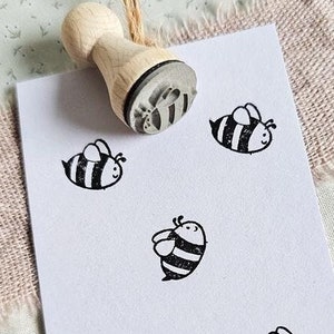 Könnte beinhalten: Eine weiße Karte mit dem Text "www.stempeldreams.de" unten gedruckt. Fünf schwarze und weiße Bienenstempel sind auf die Karte gedruckt. Ein Stempel aus Holz mit einem Bienendesign ist in der oberen linken Ecke des Bildes zu sehen.
