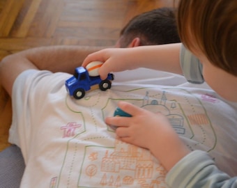 May include: A child is playing with a blue toy truck on a white t-shirt with a drawn-on road and city scene.