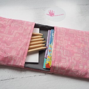 Könnte beinhalten: Rosa Stofftasche mit einem weißen geometrischen Muster. Die Tasche ist offen und enthält einen Stapel bunter Bücher, einen grauen Notizblock und einen Satz Holzstifte.