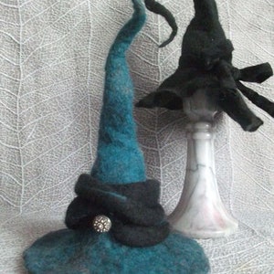 Peut inclure: Deux chapeaux de sorcière en feutre, l'un turquoise et l'autre noir. Le chapeau turquoise a une bande noire avec un bouton argenté. Le chapeau noir a un bord pointu et est sur un piédestal blanc.