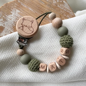 Könnte beinhalten: Ein Schnullerhalter für Babys mit einem Holzfuchs-Design. Der Clip hat Holzperlen, olivgrüne Silikonperlen und gehäkelte Perlen. Der Name "MATT" wird mit Holzbuchstabenblöcken geschrieben. Der Clip ist an einem Metallverschluss befestigt.
