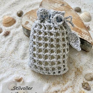 Può includere: Una sacca per sapone lavorata all'uncinetto in grigio chiaro con un motivo a griglia aperta, una chiusura a coulisse e un piccolo dettaglio decorativo a forma di foglia. La sacca contiene una saponetta e il testo "Stilvoller Seifenbeutel Häkelanleitung".
