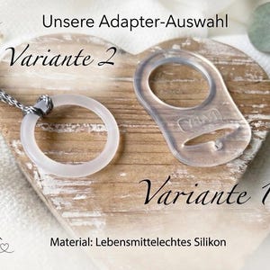 Könnte beinhalten: Zwei transparente Schnulleradapter mit der Beschriftung Variante 1 und Variante 2. Variante 1 ist ein flacher, schildförmiger Ring mit Schlitz, Variante 2 ist ein einfacher runder Ring an einer grauen geflochtenen Schnur. Text: Unsere Adapter-Auswahl.