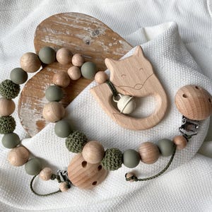 Könnte beinhalten: Ein hölzernes Baby-Beißset mit einem fuchsförmigen Beißring mit Glöckchen, einem Schnullerclip und einer Perlenkette. Die Perlen sind eine Mischung aus Naturholz, Olivgrün und gehäkelten Elementen. Das Set ist für Babys konzipiert.