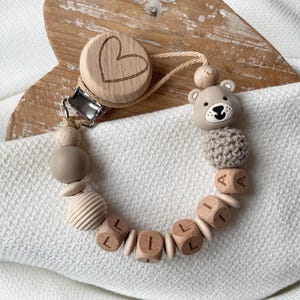 Könnte beinhalten: Ein Baby-Schnullerclip mit einem herzförmigen Holzclip und einer bärenförmigen Silikonperle. Der Clip hat Holzperlen, darunter Würfel mit dem Schriftzug "LILLA", und eine strukturierte, beige farbene Perle.