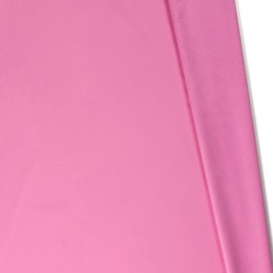 May include: A close-up of a pink fabric with a smooth texture. The fabric is folded in half, with one side lying flat and the other side folded over.