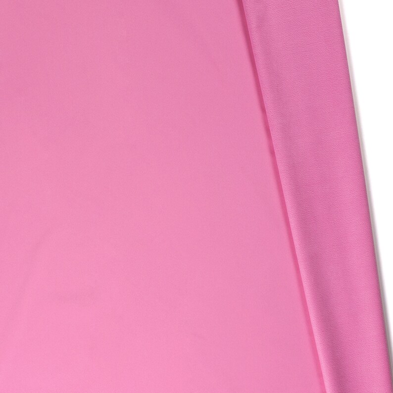 May include: A close-up of a pink fabric with a smooth texture. The fabric is folded in half, with one side lying flat and the other side folded over.
