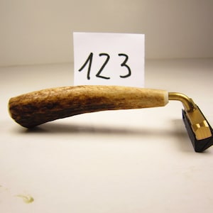 Könnte beinhalten: Ein Rasierer mit einem strukturierten, braun-cremefarbenen Geweihgriff und einem goldfarbenen Metallkopf mit schwarzer Klingenpatrone. Hinter dem Griff befindet sich eine weiße quadratische Karte mit der handgeschriebenen schwarzen Zahl 123.