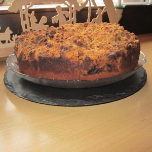 Puede incluir: Un pastel redondo con una cobertura de migas, cortado por el centro, se asienta sobre un plato de cristal transparente sobre una pizarra negra. El pastel tiene una corteza marrón dorada y un interior marrón oscuro.