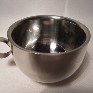 Peut inclure: Une tasse en acier inoxydable brillante, de forme arrondie avec une anse circulaire. La surface de la tasse est réfléchissante et semble vide. L'anse est fixée sur le côté de la tasse.