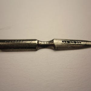 Könnte beinhalten: Eine Vintage-Metall-Kalligraphie-Feder mit spitzer Spitze und dekorativen Aussparungen. Die Feder hat gravierten Text auf dem Schaft und ist für detailliertes Schreiben oder Zeichnen konzipiert.