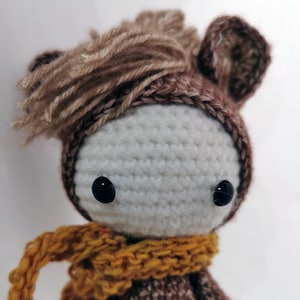 Könnte beinhalten: Ein gehäkeltes Stofftier aus braunem und weißem Garn, das einen braunen Hut mit einem braunen Garnpompon und einen gelben Schal trägt.