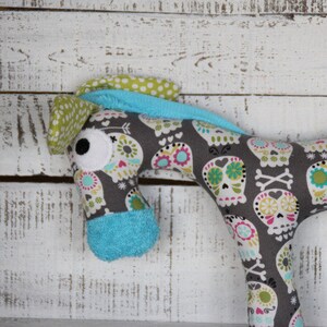 以下が含まれることがあります： 骸骨と花の模様がついたグレーとブルーのぬいぐるみ馬。馬は青と白のドットの鼻と大きな白い目を持っている。